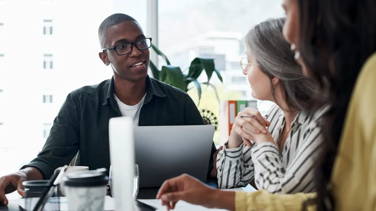 Group of colleagues having a discussion in a modern office, with one person speaking while using a laptop during a meeting.